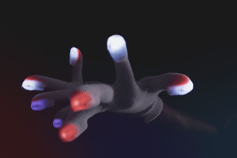 Gloved hand in a claw shape, fingertips lit in red, purple, and white against a dark background